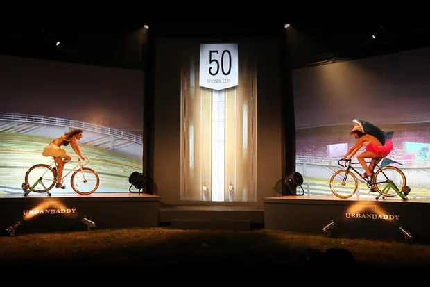 Two women riding bikes on a stage with projection backgrounds.